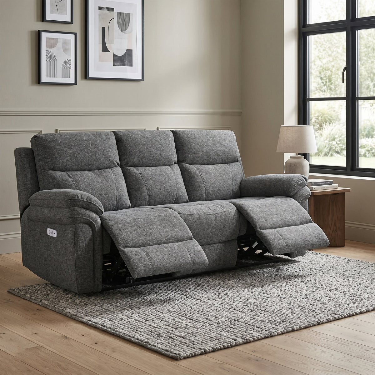 A gray fabric reclining loveseat sofa shown in a staged living room with a window, side table, lamp, wall art, rug, and wood floor.