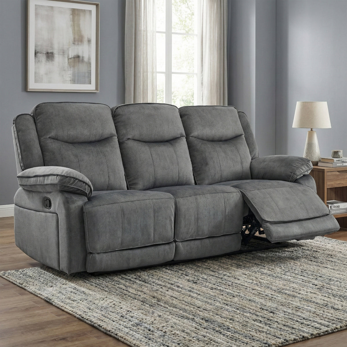 A three-seat gray reclining sofa with one cushion pulled out on a patterned area rug in a living room setting.