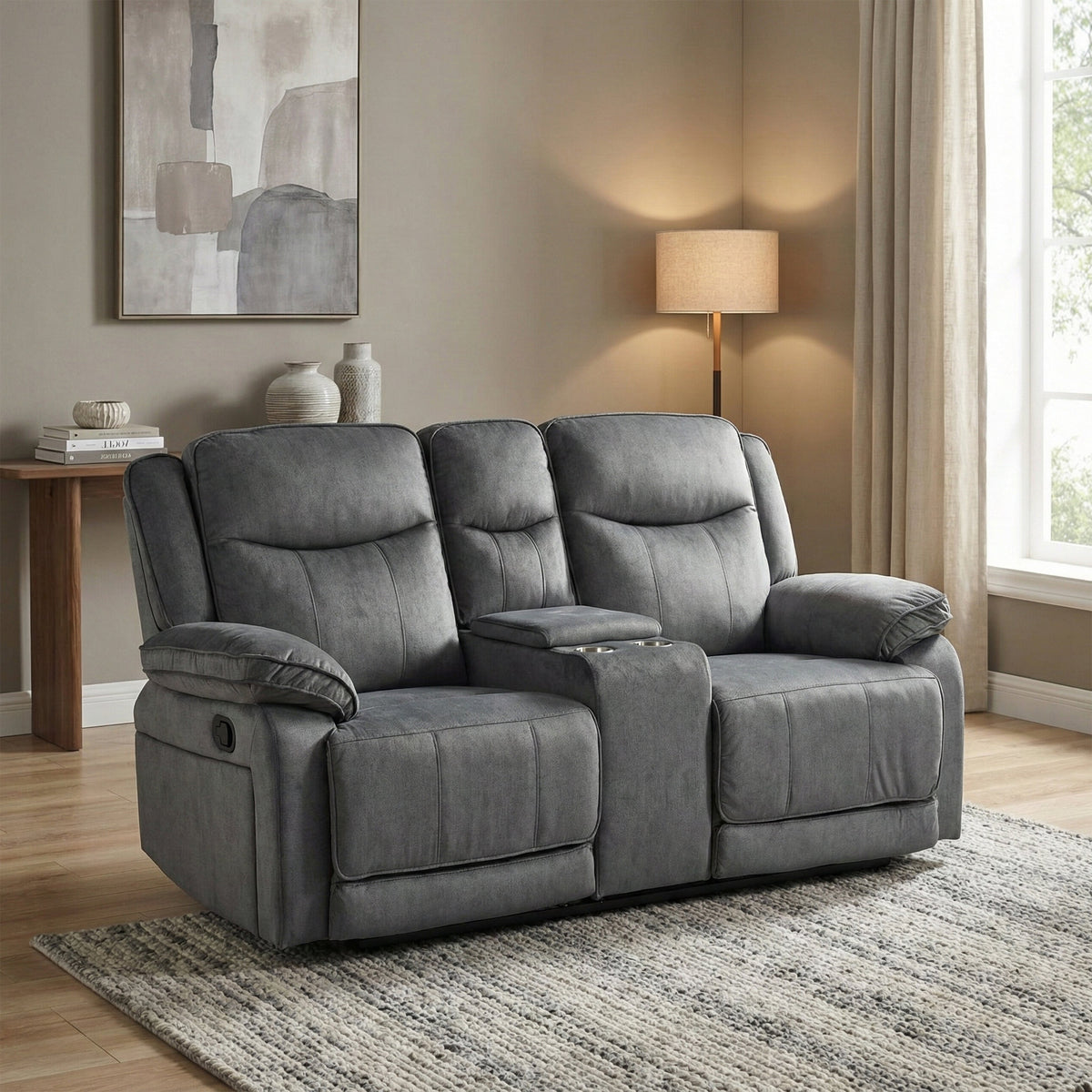 A gray fabric reclining loveseat with two seat cushions, wide armrests, and a center console cup holder, shown in a staged living room setting with a lamp, side table, curtains, and rug.
