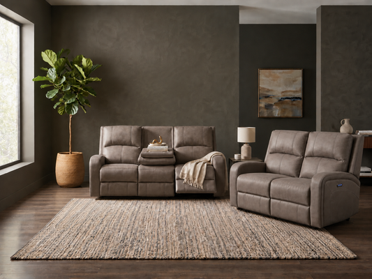 A living room scene showing a taupe-brown reclining loveseat and a matching reclining armchair on a large woven area rug, with dark gray walls, wood flooring, a potted plant, and wall art in the background.